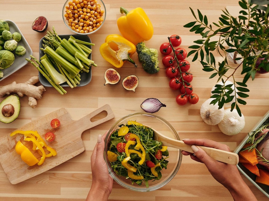 A person stirs a salad on a bench covered with fresh vegetables and legumes.