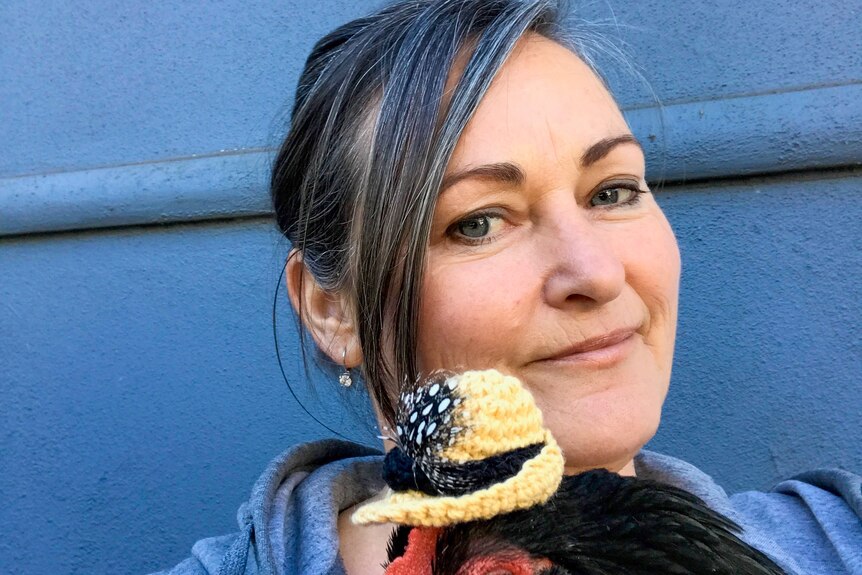 Mandy Watts is seen in a selfie smiling as she holds a chicken wearing one of her crochet hat creations.