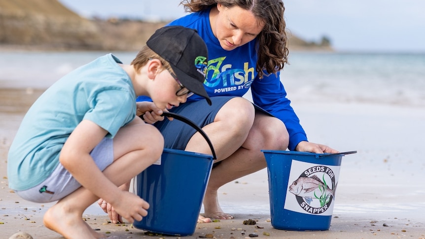 A woman and young boy holding buckets and kneeling on the beach.