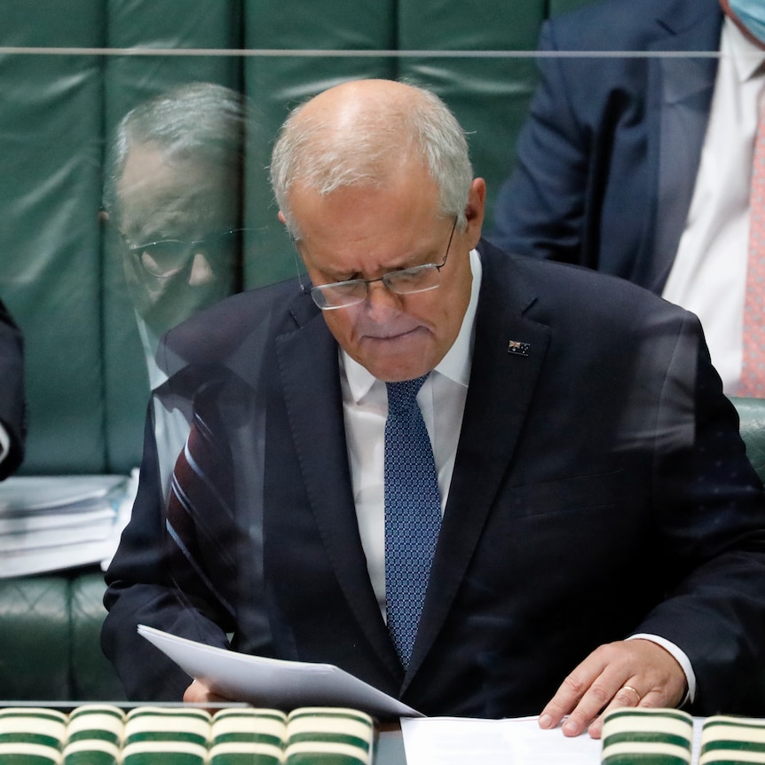 Scott Morrison looks down at papers. Anthony Albanese is in the reflection of a protective shield in front of Morrison