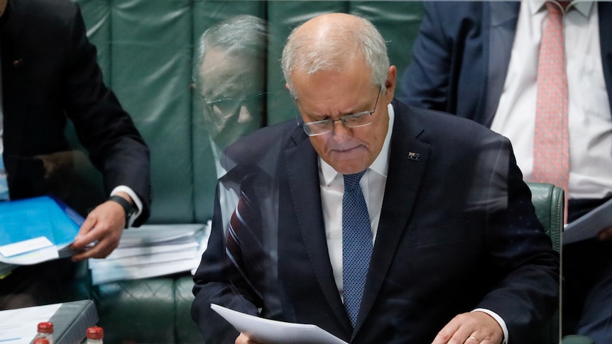 Scott Morrison looks down at papers. Anthony Albanese is in the reflection of a protective shield in front of Morrison