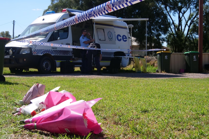 Floral bouquets lie on a lawn near a police van parked in a driveway.