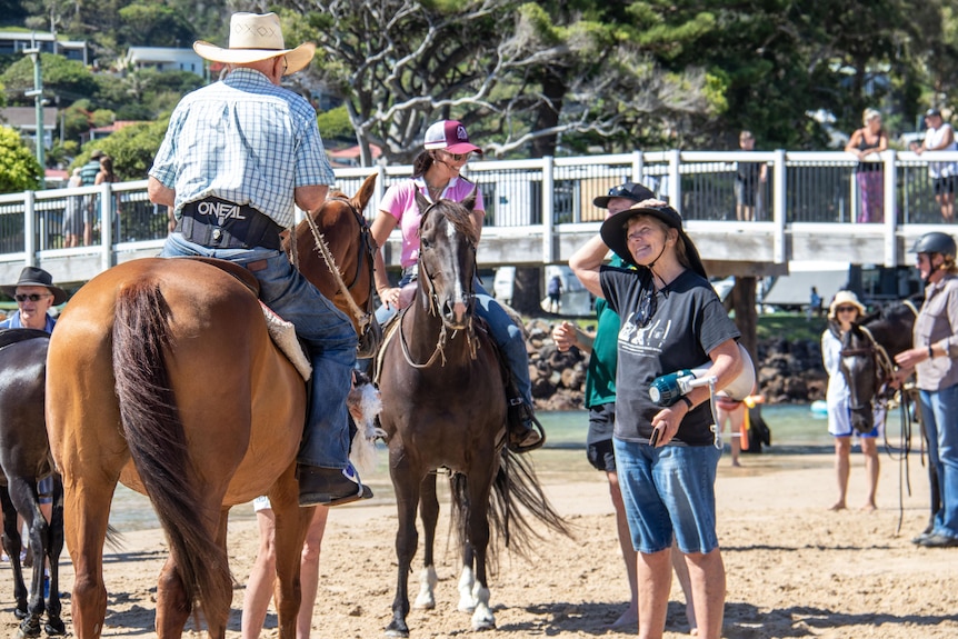 Woman with jean shorts and black shirt stands talking to two people on horseback, standing on a beach