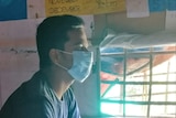 A man sitting in a room with posters, wearing a face mask