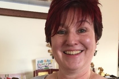 A head and shoulders shot of a woman with cropped dark red hair stands in a living room.