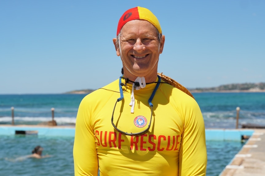 A man wearing a surf rescue rash vest and cap stands at the beach smiling.
