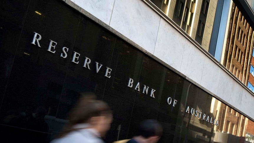 Two people blur as they walk past a sign on a black tiled wall. It says RESERVE BANK OF AUSTRALIA