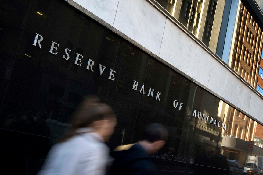 Two people blur as they walk past a sign on a black tiled wall. It says RESERVE BANK OF AUSTRALIA