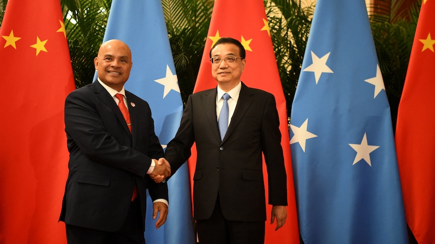 Two men in suits smile as they shake hands before the flags of Micronesia and China at an official event.