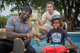 Two men and a child smile at the camera as they sit on top of a skateboarding ramp after a community skateboarding workshop.