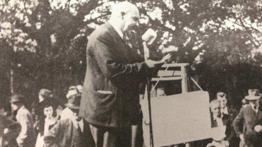 A black and white photo of a man giving a speech into a microphone in front of a crowd in the street.