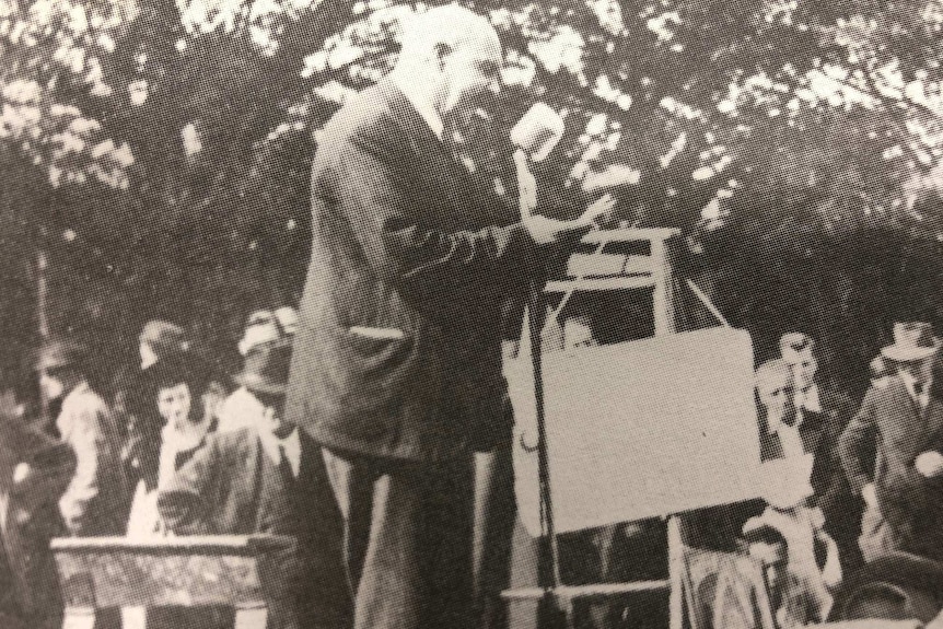 A black and white photo of a man giving a speech into a microphone in front of a crowd in the street.