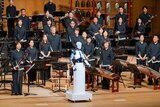White humanoid robot EveR 6 stands at the front of the stage in front of standing members of the orchestra facing the audience