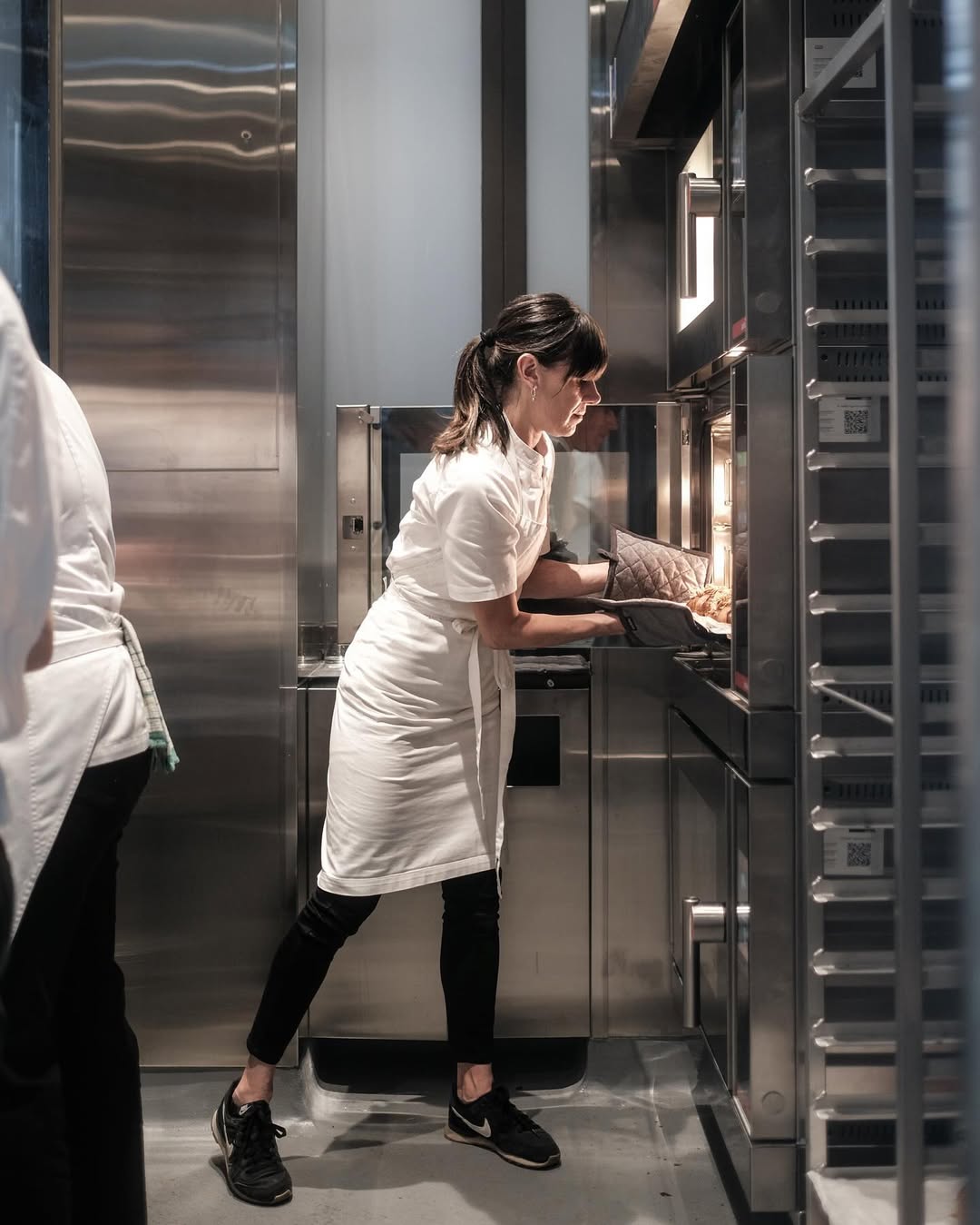 A woman with brown hair wears a chef's white shirt and long pants putting a tray of croissants in the oven