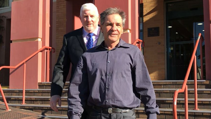 A man in a stripey purple and black shirt walking out of a court  building with a lawyer in a suit behind him