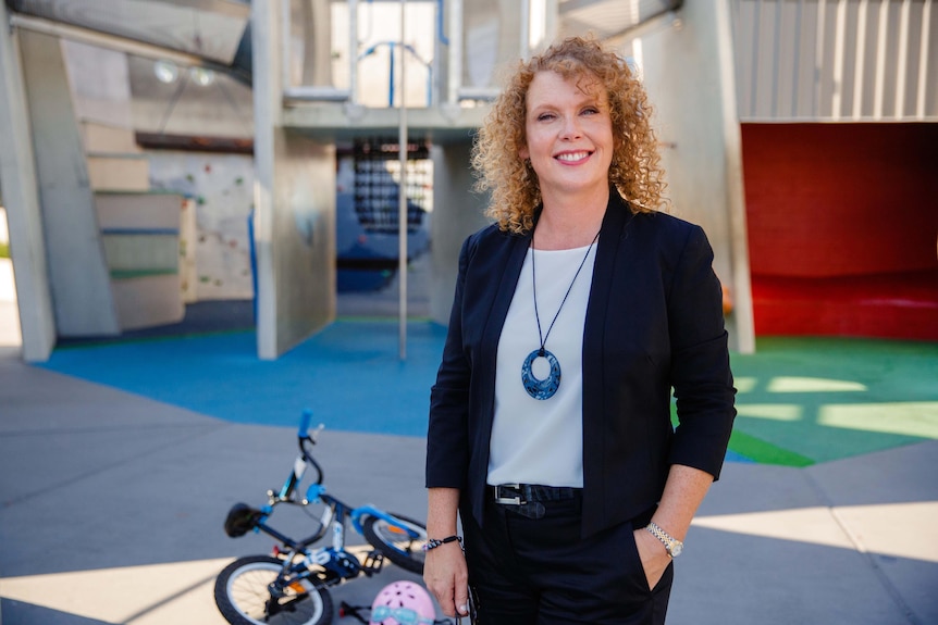 Karen Barlow, who has copper curly hair and blue eyes, poses for a photo outside, a child's bike on the ground next to her