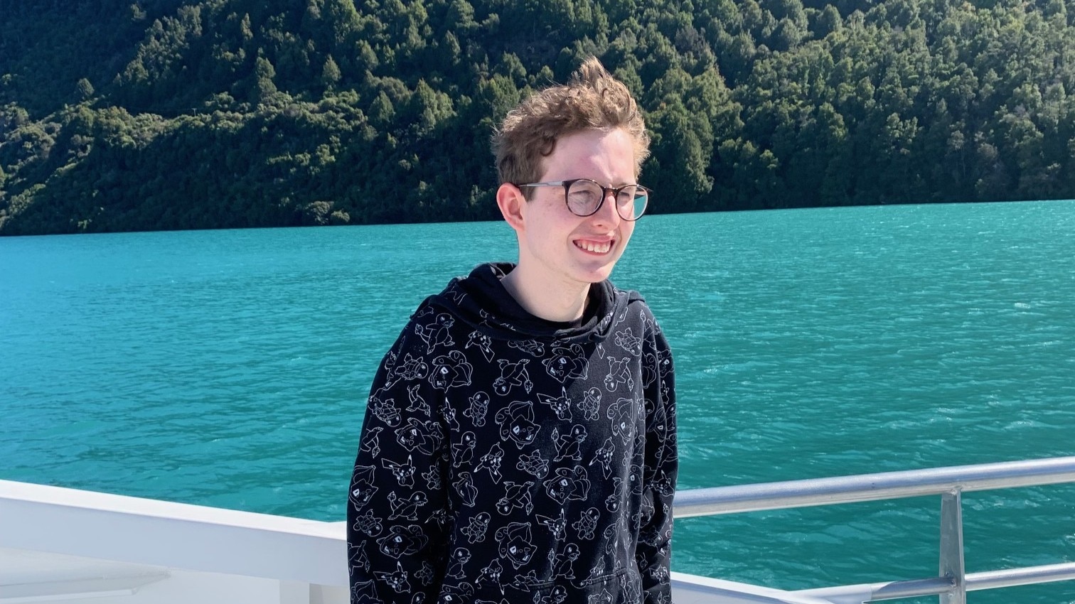 A young man with glasses standing on a boat. Behind him is a large mountain and water. 