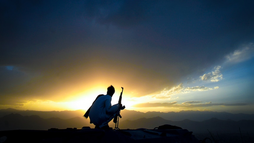 A soldier crouching next to a rifle looking at the sun setting behind the hills on the horizon