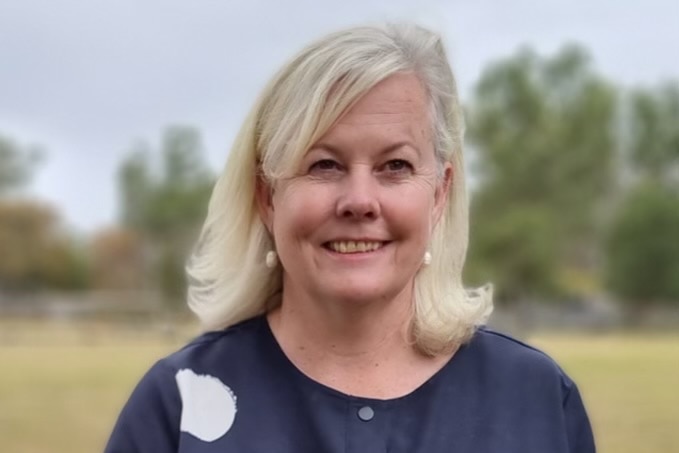 A smiling, middle-aged woman with blonde hair stands in a paddock.