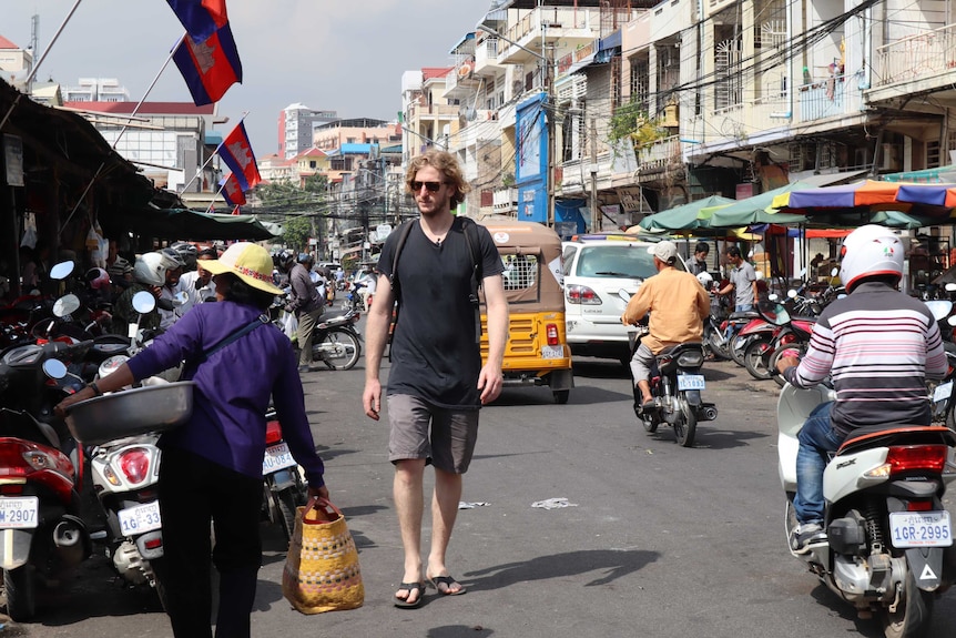 A man wearing sunglasses, tshirt, shorts and a backpack walks through a Cambodia street filled with vehicles