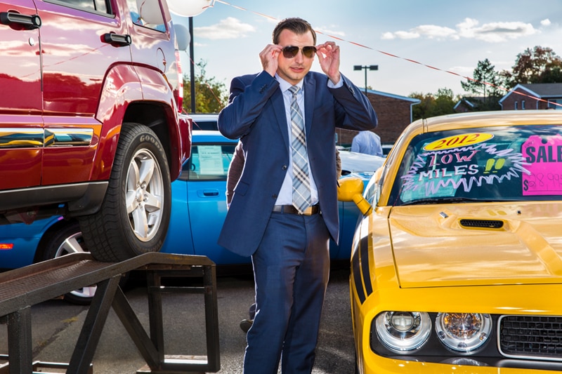 Sales consultant Jason Mascia in a blue suit adjusts his sunglasses in between two Jeeps for sale.