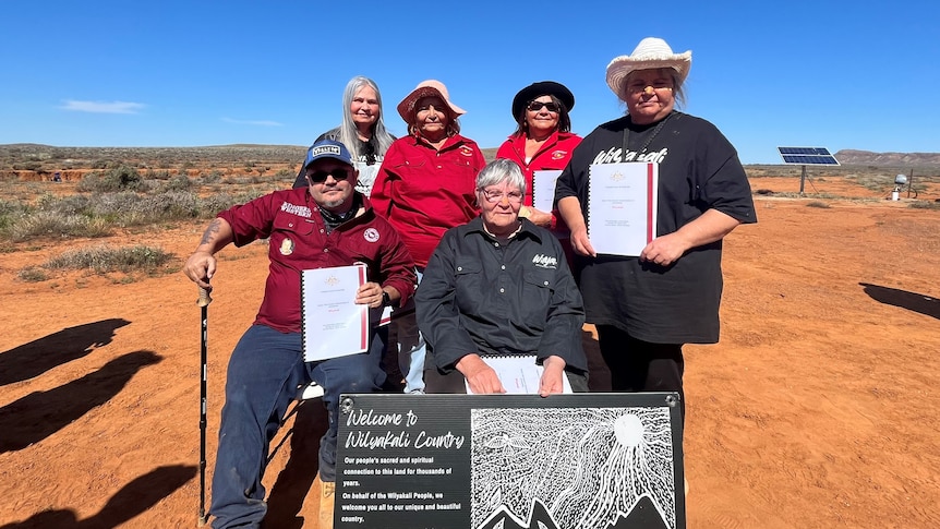 A group of Aboriginal men and women standing and sitting on a red patch of dirt in front of a small sign