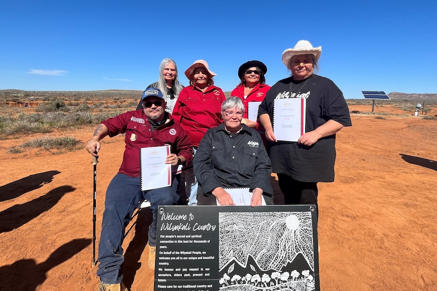 A group of aboriginal men and women standing and sitting on a red patch of dirt in front of a small sign