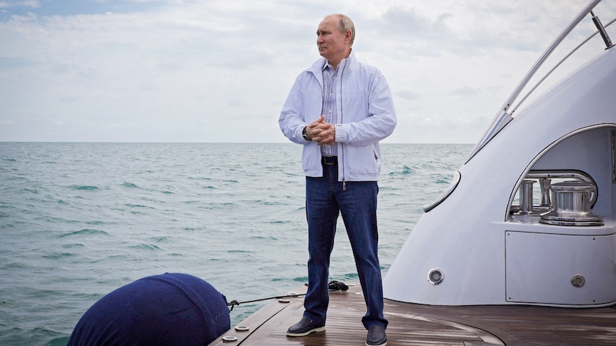 Vladimir Putin in a white jacket and jeans stands on the back of a yacht looking out to sea