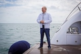 Vladimir Putin in a white jacket and jeans stands on the back of a yacht looking out to sea