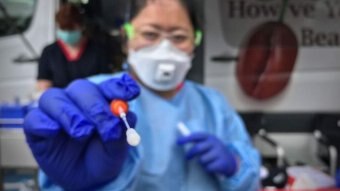 Health worker with PPE holds a testing swab toward the camera.