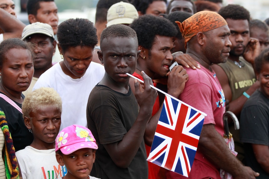 A boy holds a union flag in his finger, resting it on his lip, among a crowd