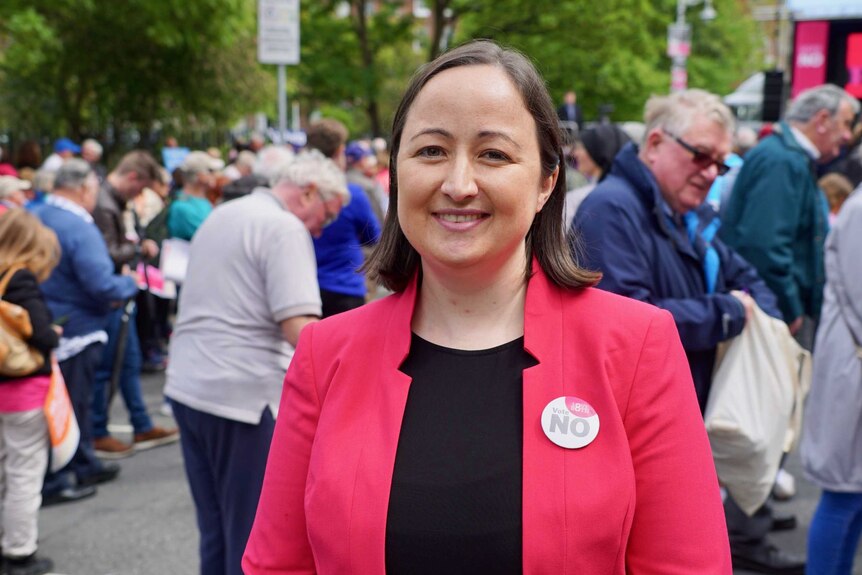 A woman with shoulder-length brown hair wears a coral red jacket with a pin that reads 'Vote NO'