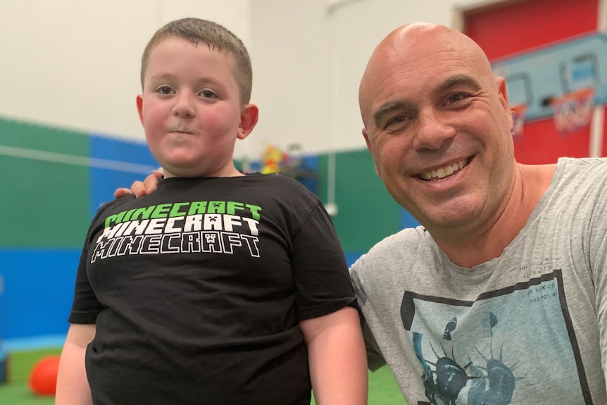 Max Drew stands on an indoor sports court with his sports therapist Jason Nanos kneeling beside him