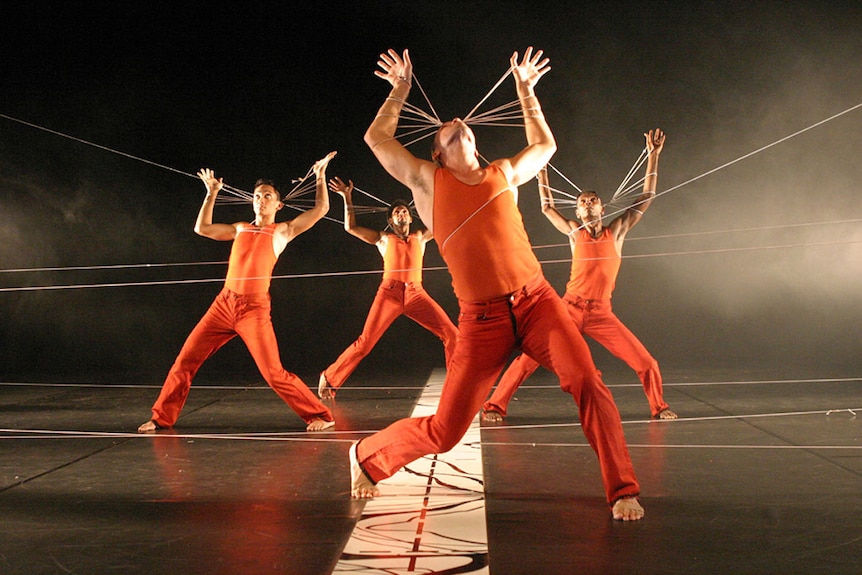 Four male dancers in red costumes perform on stage with raised arms bound together by string.