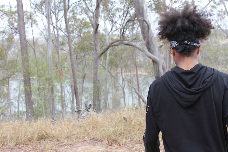 A young man with curly black hair stands overlooking trees and a river.