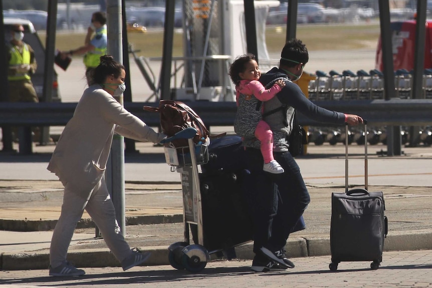A man and a woman with a suitcase and a luggage trolley and a child on the man's back walk outside Perth Airport.