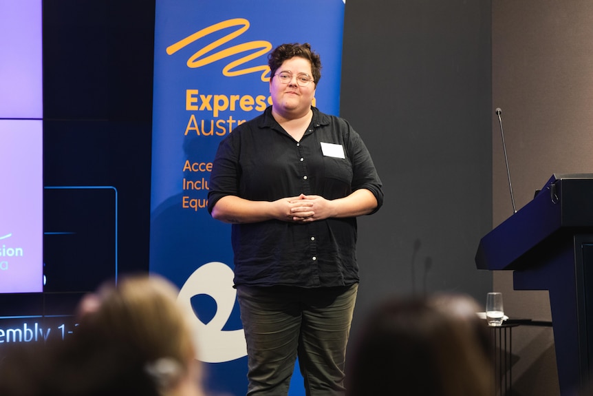 Sherrie Beaver in a black shirt stands before an audience and next to a podium. Behind her is a banner for Expression Australia.