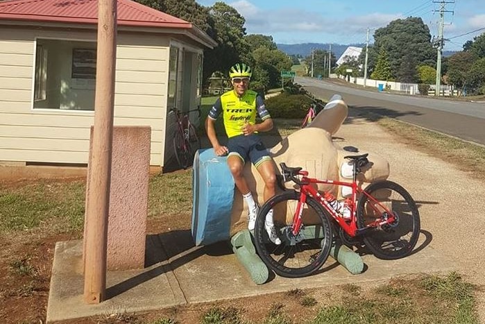 Cyclist Richie Porte sits on a giant thumb holding his own thumb up.