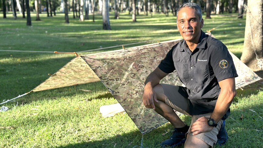 A man with brown skin, dark shirt and grey hair at a park near trees and green grass.