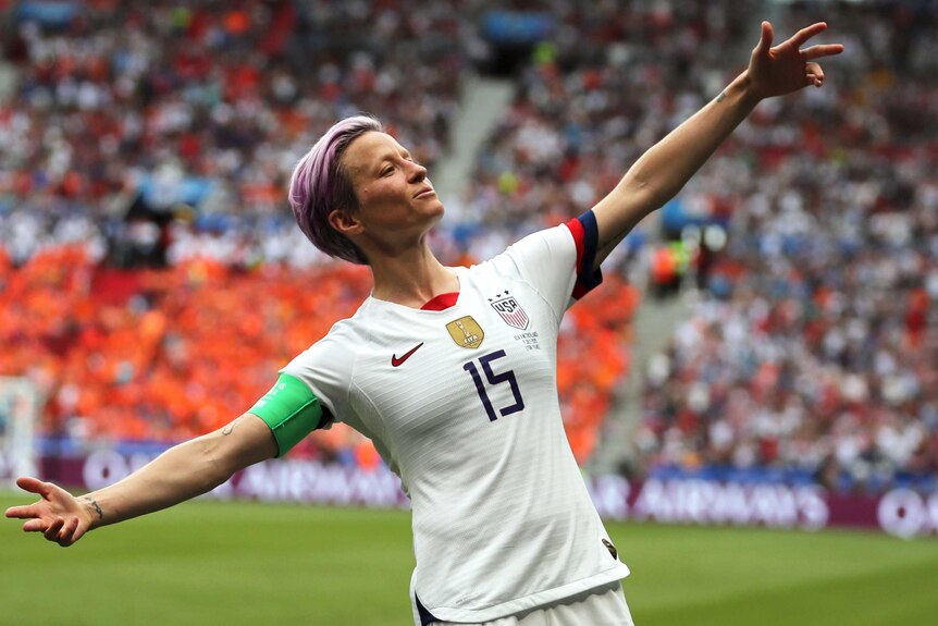 Megan Rapinoe stands with her arms outstretched in front of a crowd of spectators