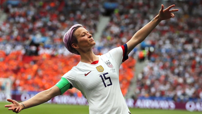 Megan Rapinoe stands with her arms outstretched in front of a crowd of spectators