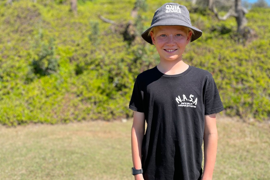 A 12-year-old boy in a bucket hat and his surf boardriders club t-shirt