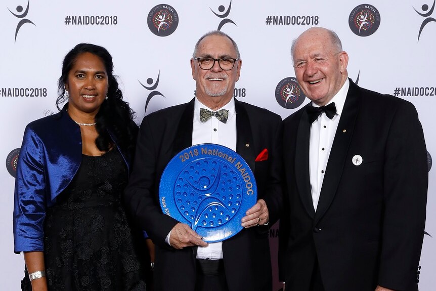 Russell Taylor holds a large, round, blue Naidoc award,standing between a woman and a man, all on a red carpet