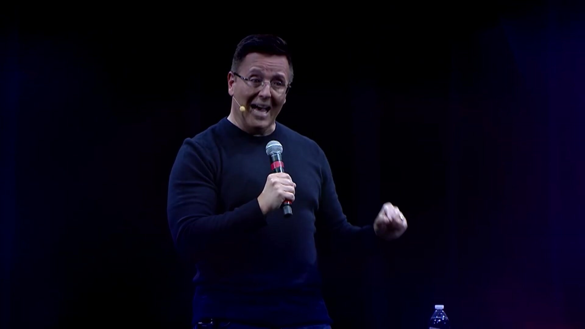 TV psychic John Edward stands in a dark room, holding a microphone, speaking to a crowd. He wears glasses and a dark jumper.