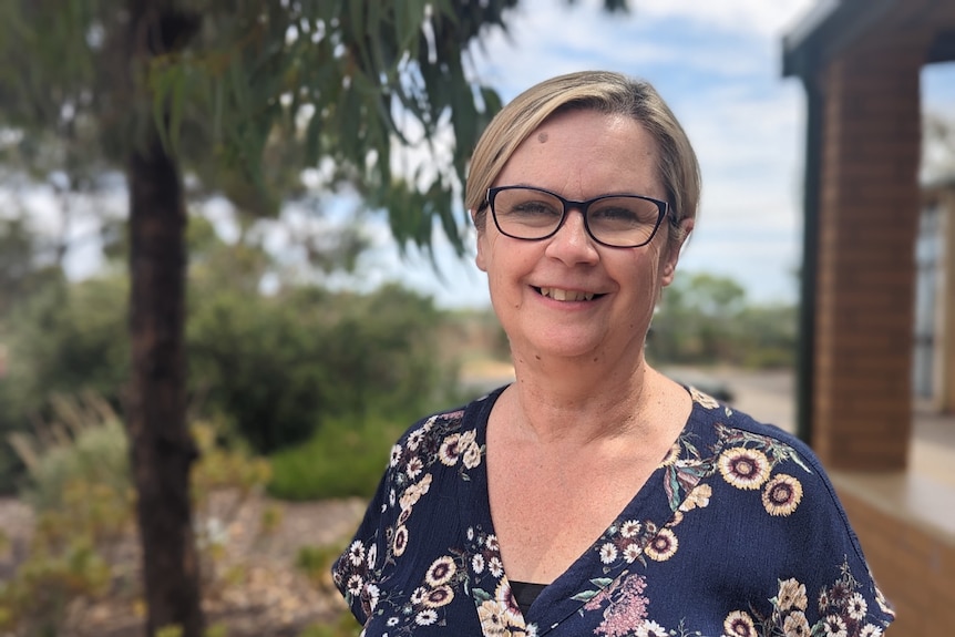 A white woman, Clare Scriven, with short blonde hair and glasses and a blue floral dress smiling at the camera.