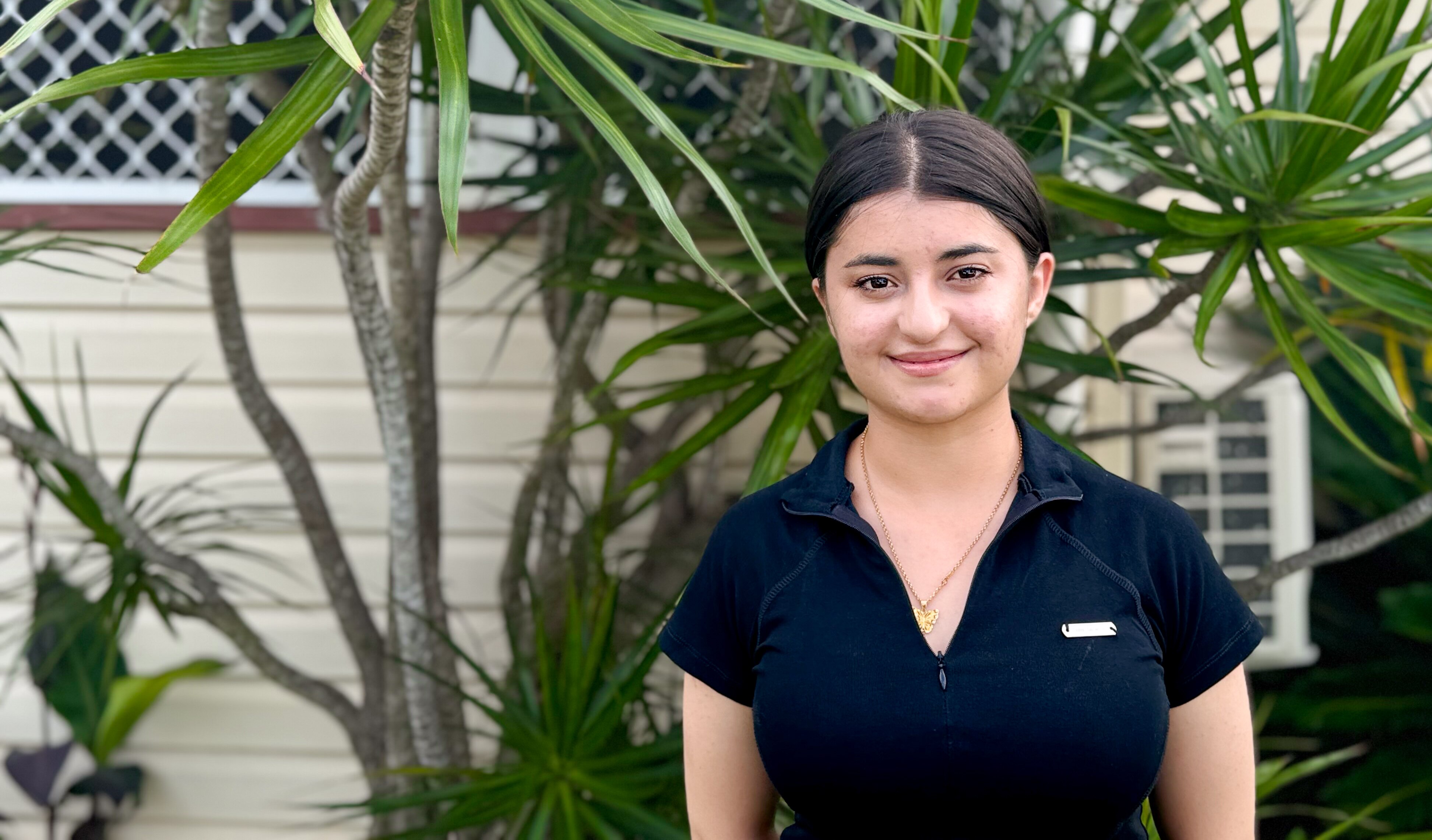 A young woman with light olive skin and black hair smiles at the camera in front of a house and green plants