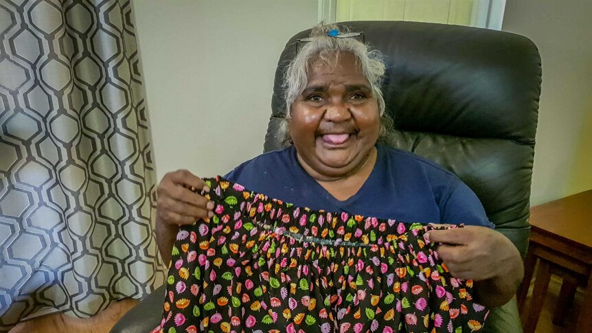 An Indigenous woman with silver hair sits in an armchair holding up a colourful skirt.