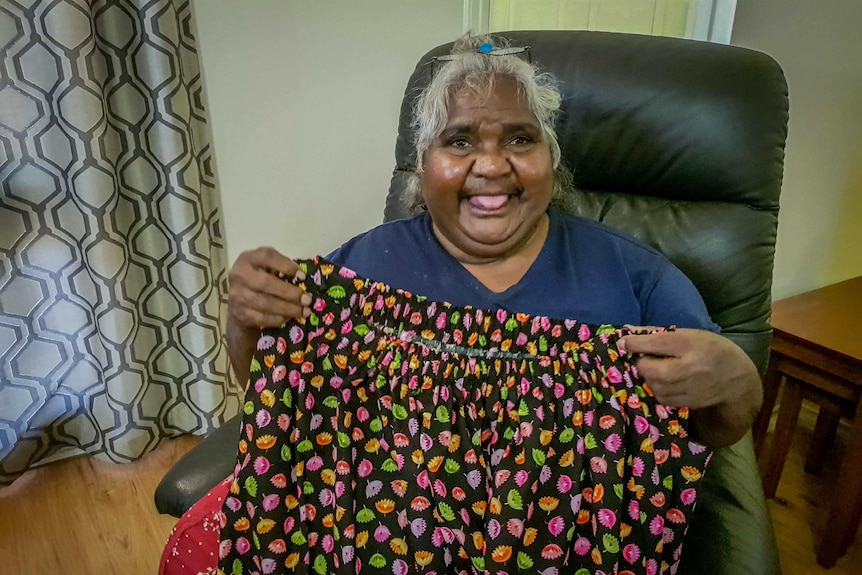An Indigenous woman with silver hair sits in an armchair holding up a colourful skirt.