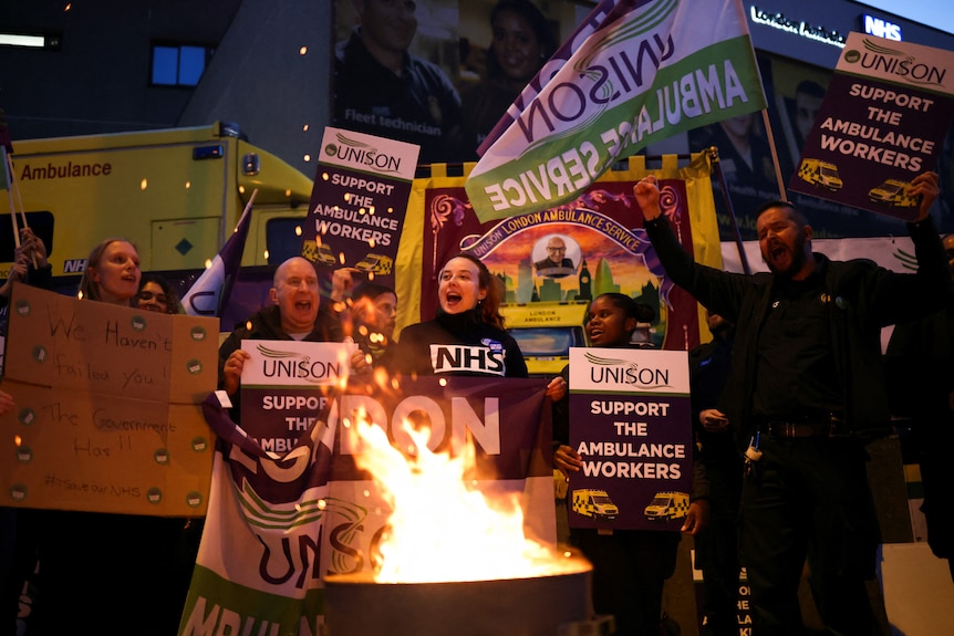 A group of people holding signs gather by a fire on a road with ambulances lined up behind them.