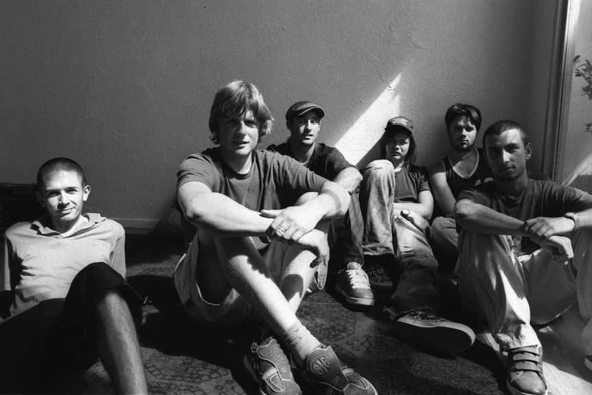 Black and white image of six young Cat Empire band members sitting on the floor with arms crossed over their knees. 
